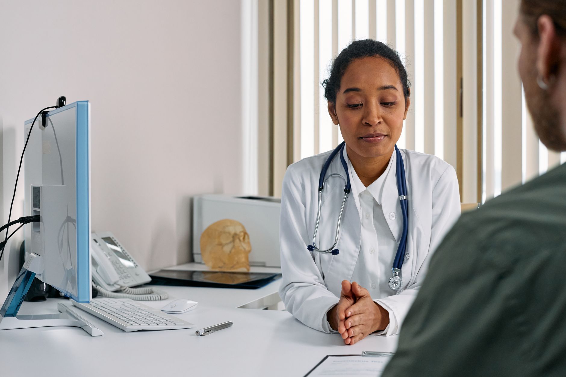 Doctor in white coat consults patient at desk with computer monitor and stethoscope, clinical office setting.