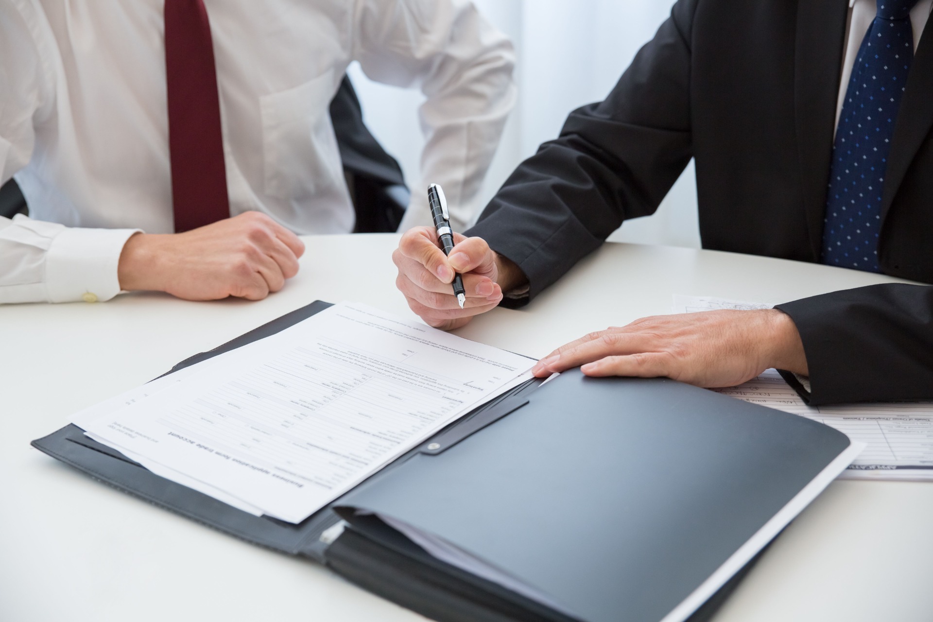 Two people in suits review documents, one holding a pen and signing paperwork at a table.