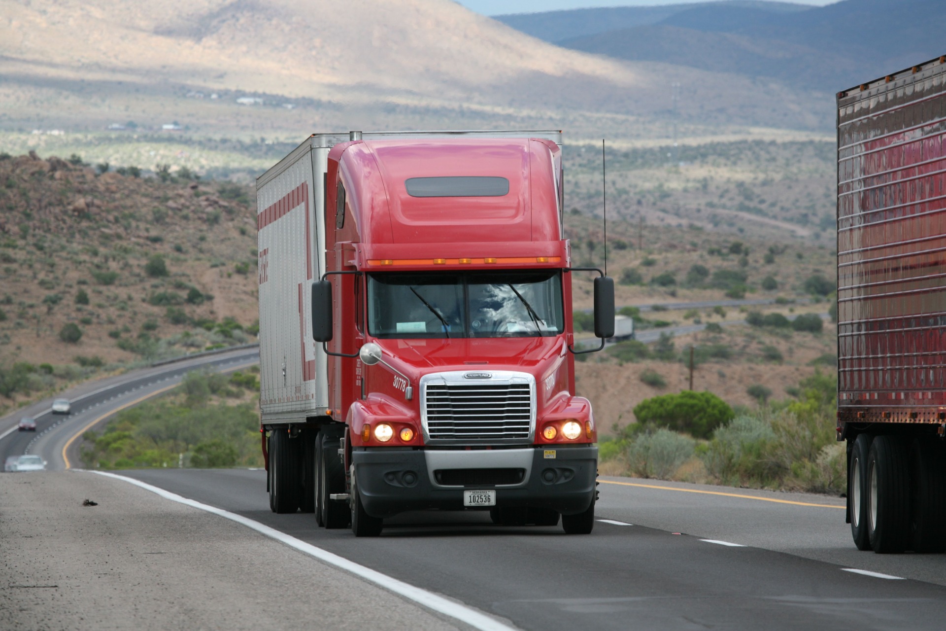 Red semi truck with cargo trailer drives on highway through mountainous landscape, viewed from front.