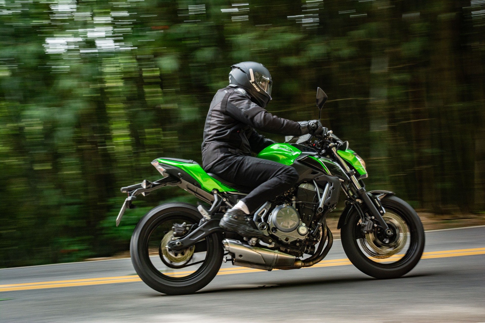 Motorcyclist in black gear rides a bright green sport bike on a road, blurred trees behind.