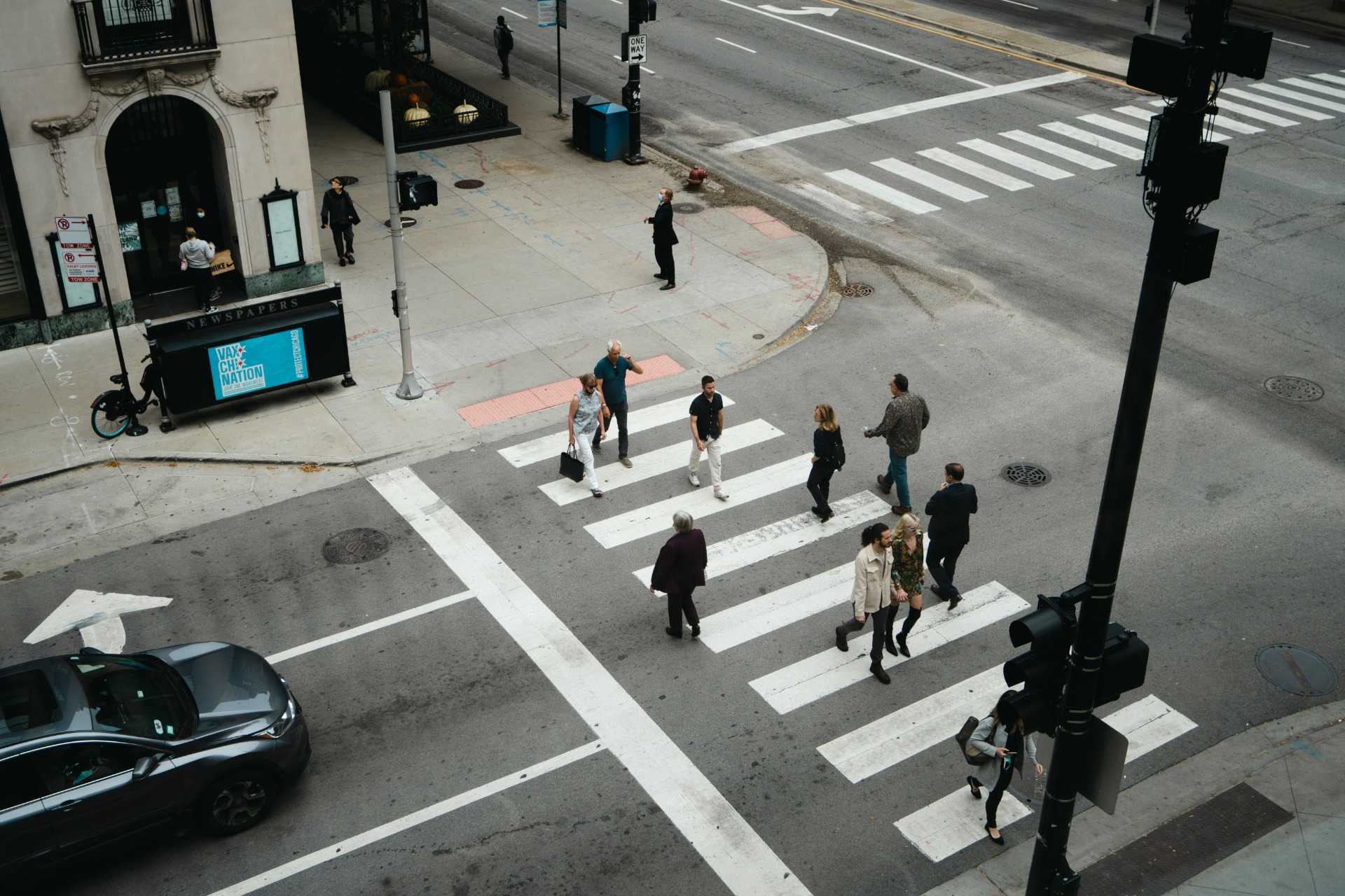 People cross a city street at a marked crosswalk near storefronts and traffic lights.