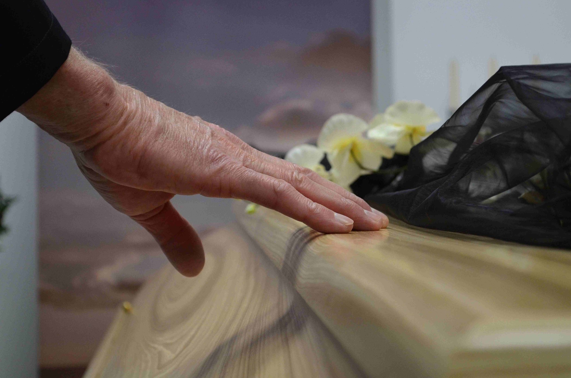 Elderly hand gently smoothing a wooden coffin, with blurred yellow flowers in the background.