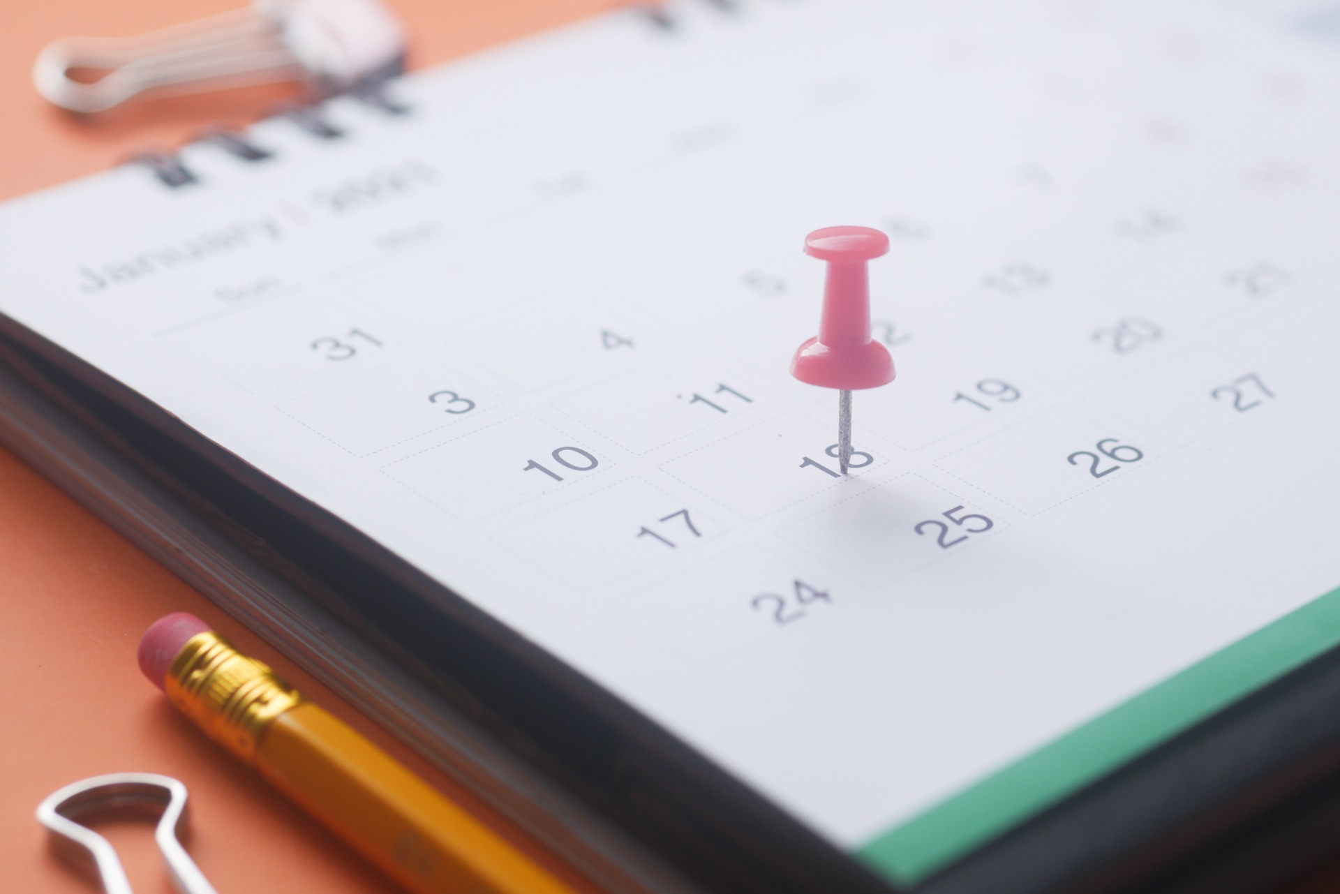 Pink pushpin marks a calendar date, with numbers visible and stationery items blurred in foreground.