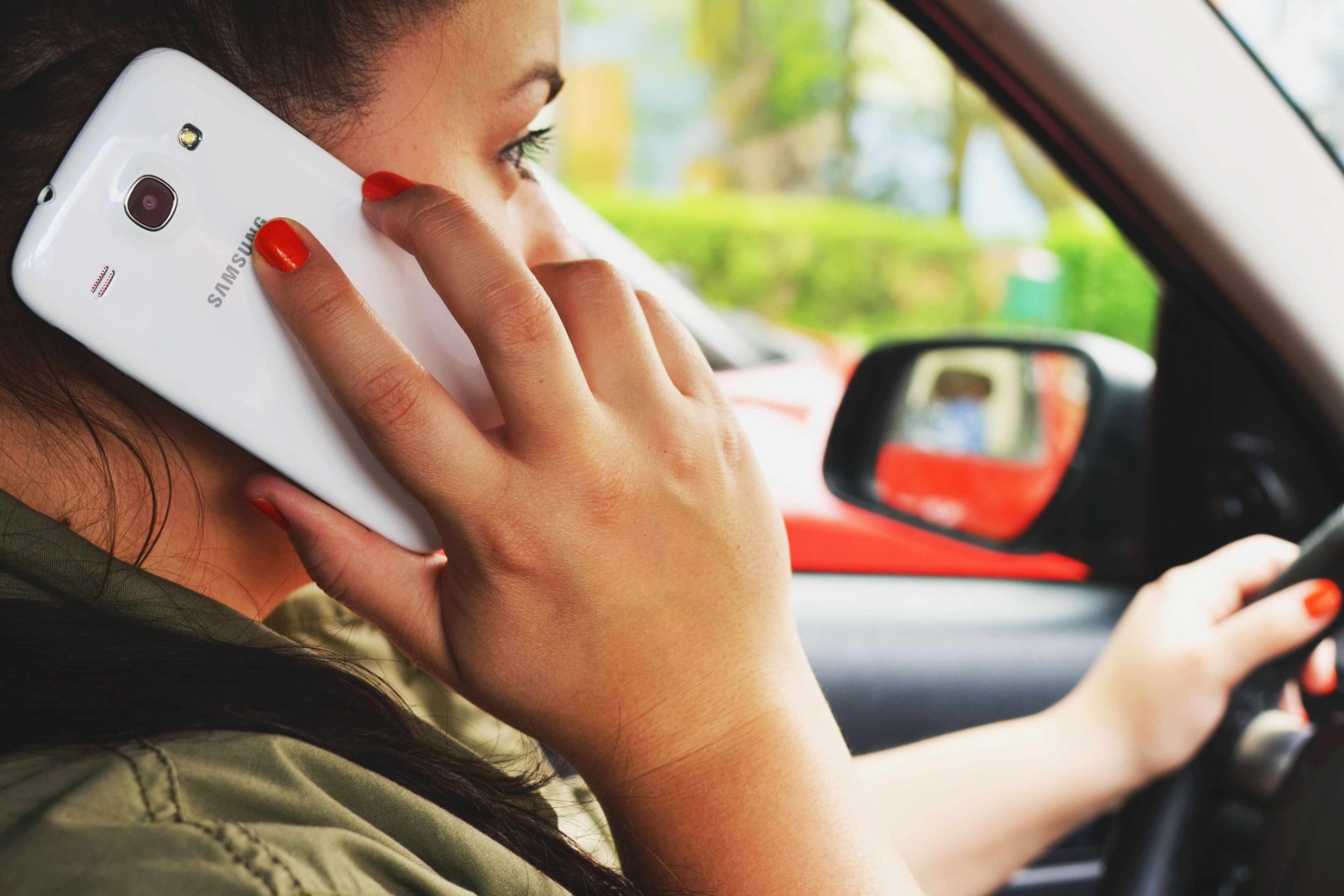 Woman driving with hand on steering wheel, holding smartphone to ear, red nails, sunny blurred background.