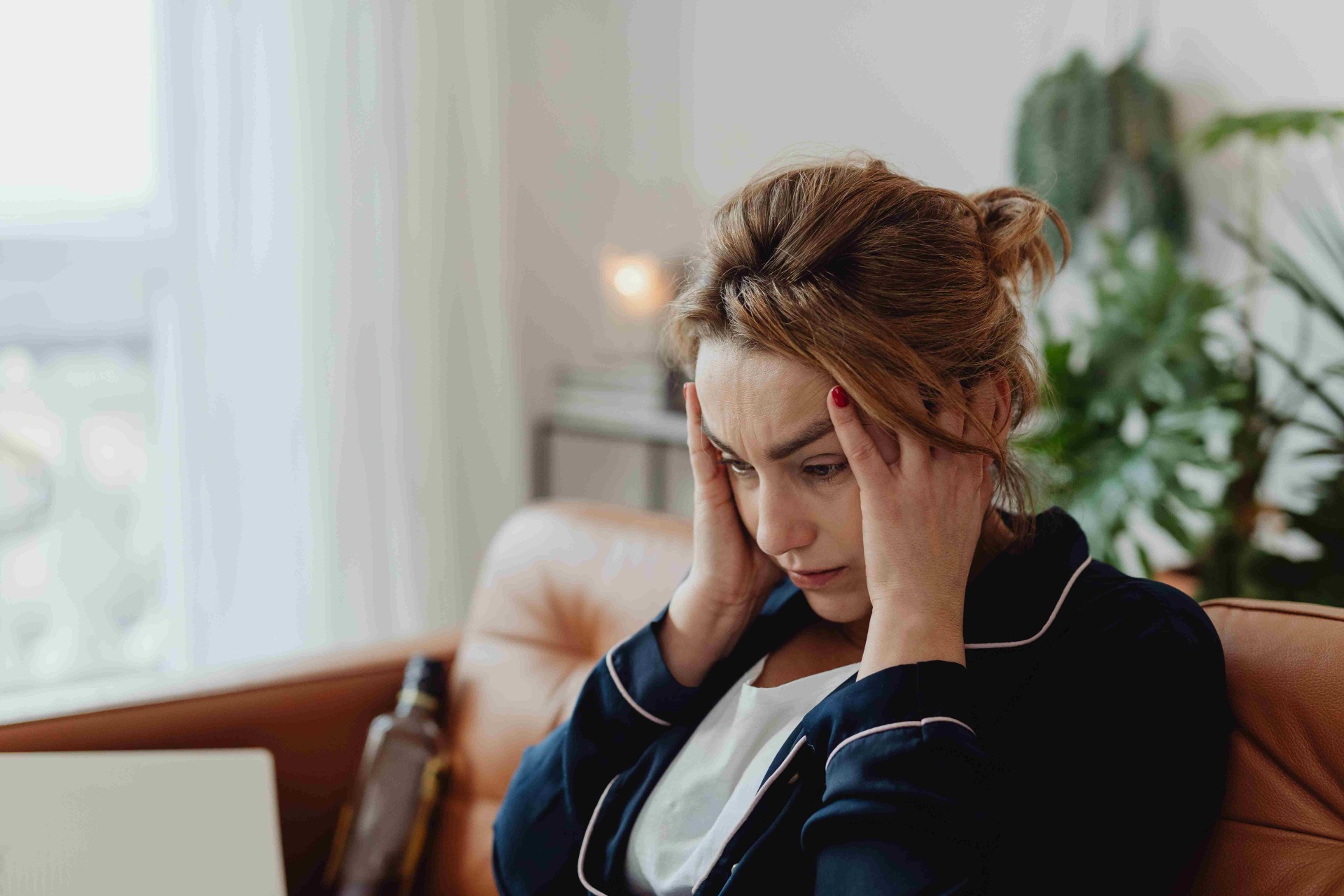 Woman with red hair in pajamas sits on couch, hands on temples, looking stressed indoors.