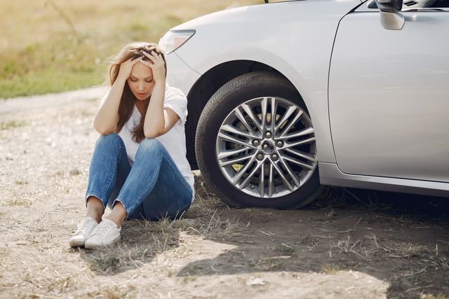 Woman sits beside a car, holding her head, with one hand on her forehead near the tire.